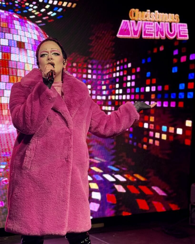 Eine Sängerin mit Mikrofon, bekleidet mit einem pinken Mantel, tritt bei der Christmas Avenue auf.