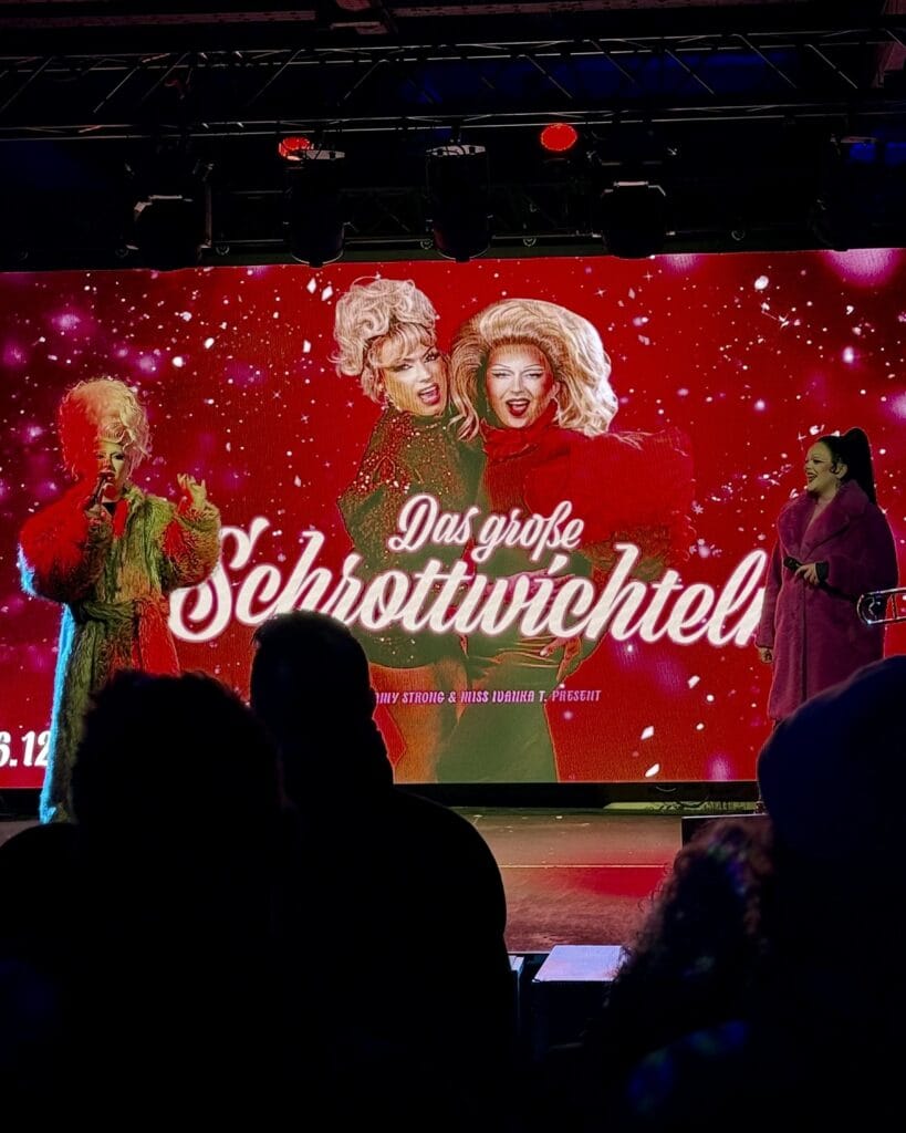 Besucher sehen eine Bühnenshow namens "Das große Schrottwichteln" mit drei Dragqueens in glamourösen Outfits vor einem roten Hintergrund.