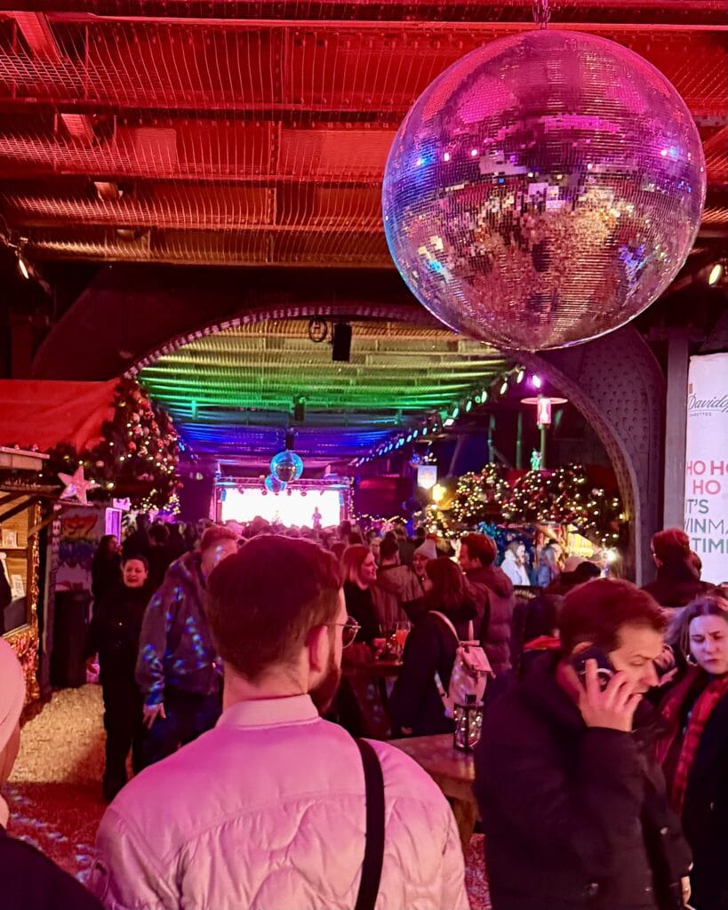 Eine glitzernde Spiegelkugel hängt über einer Menschenmenge auf der Christmas Avenue, die eine festliche Atmosphäre mit Musik genießt.