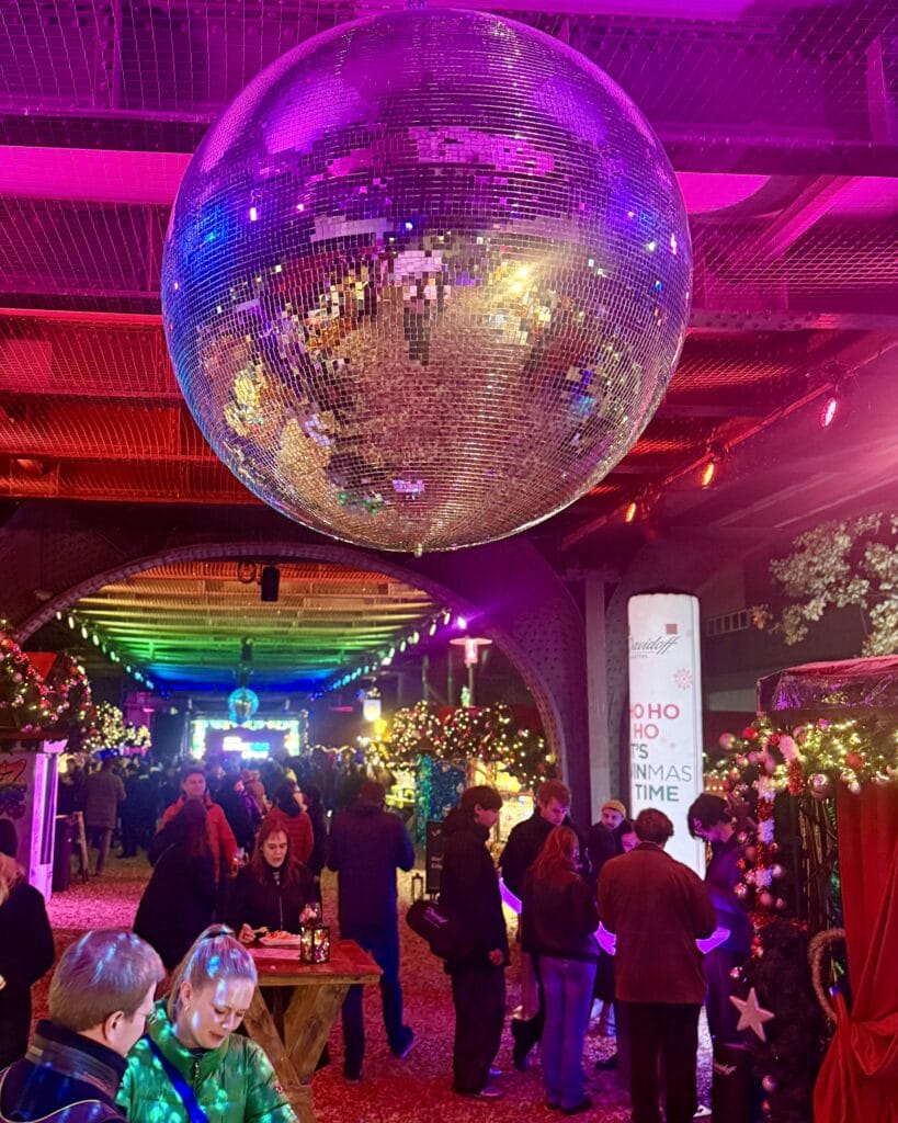 Auf dem Weihnachtsmarkt unter einer Eisenbahnbrücke hängt eine große Discokugel über den Besuchern, die an Tischen stehen.
