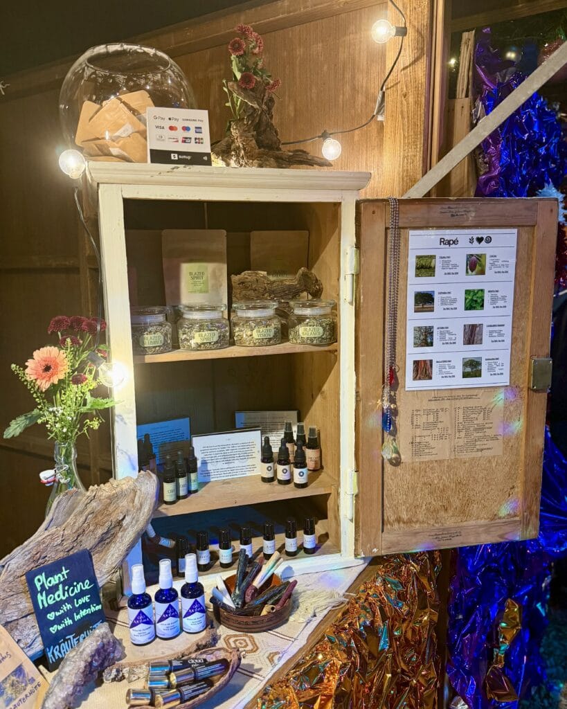 Ein offener, weißer Schrank dient als Verkaufsstand und präsentiert Kräuter, Öle und andere Naturprodukte, stimmungsvoll beleuchtet durch Lichterketten.