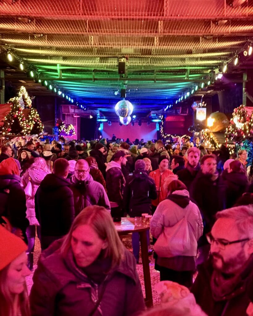 Besucher genießen die festliche Atmosphäre auf einem Weihnachtsmarkt mit bunten Lichtern, Bühne und geschmückten Weihnachtsbäumen.