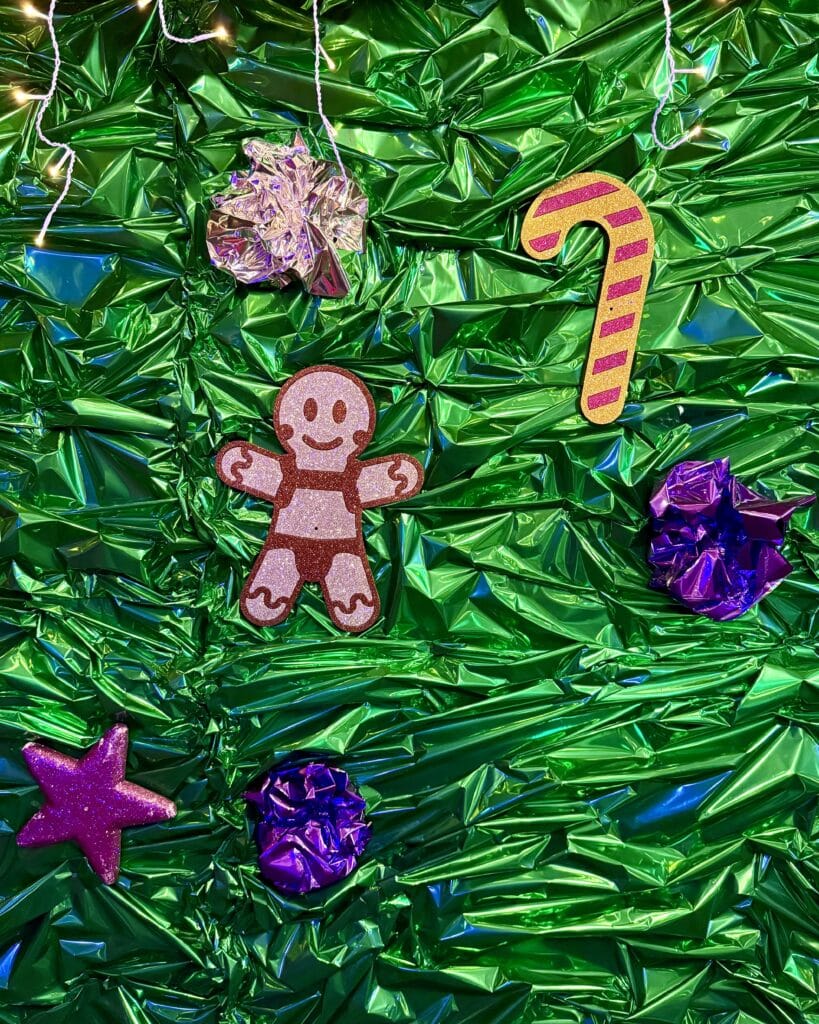 Vor einem glänzend grünen Hintergrund hängen verschiedene Schmuckstücke wie ein Lebkuchenmann, eine Zuckerstange und kleine, glitzernde Sterne.