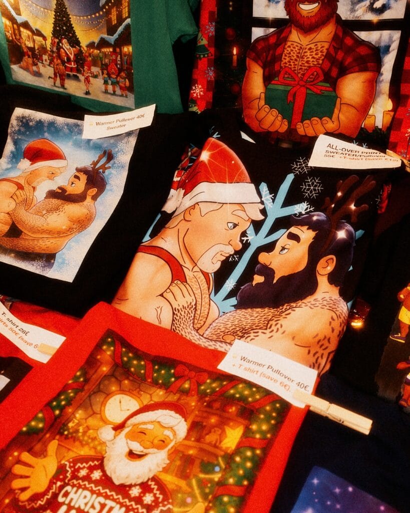 Auf einem Verkaufsstand liegen T-Shirts mit weihnachtlichen Motiven und muskulösen Männern, mit Preisschildern und Wäscheklammern befestigt.