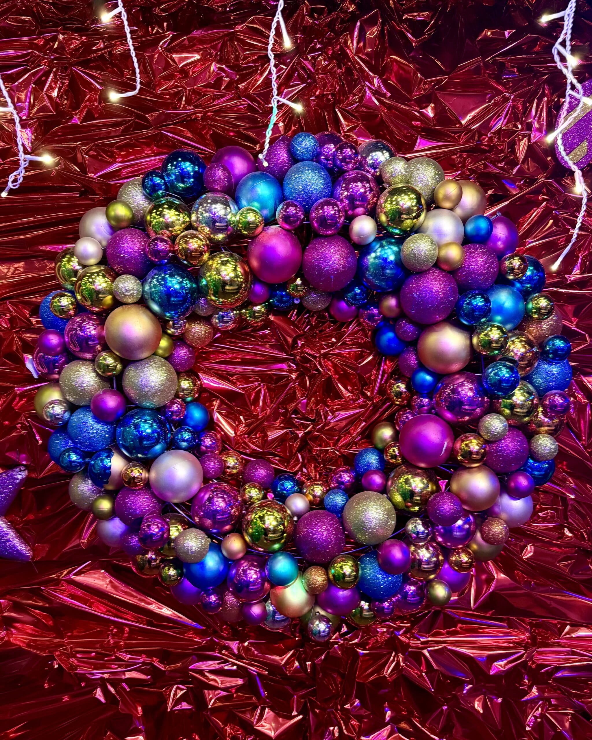 Ein farbenfroher Weihnachtskranz, geschmückt mit glitzernden Kugeln in Violett, Blau und Gold, hängt vor rotem, glänzendem Hintergrund.
