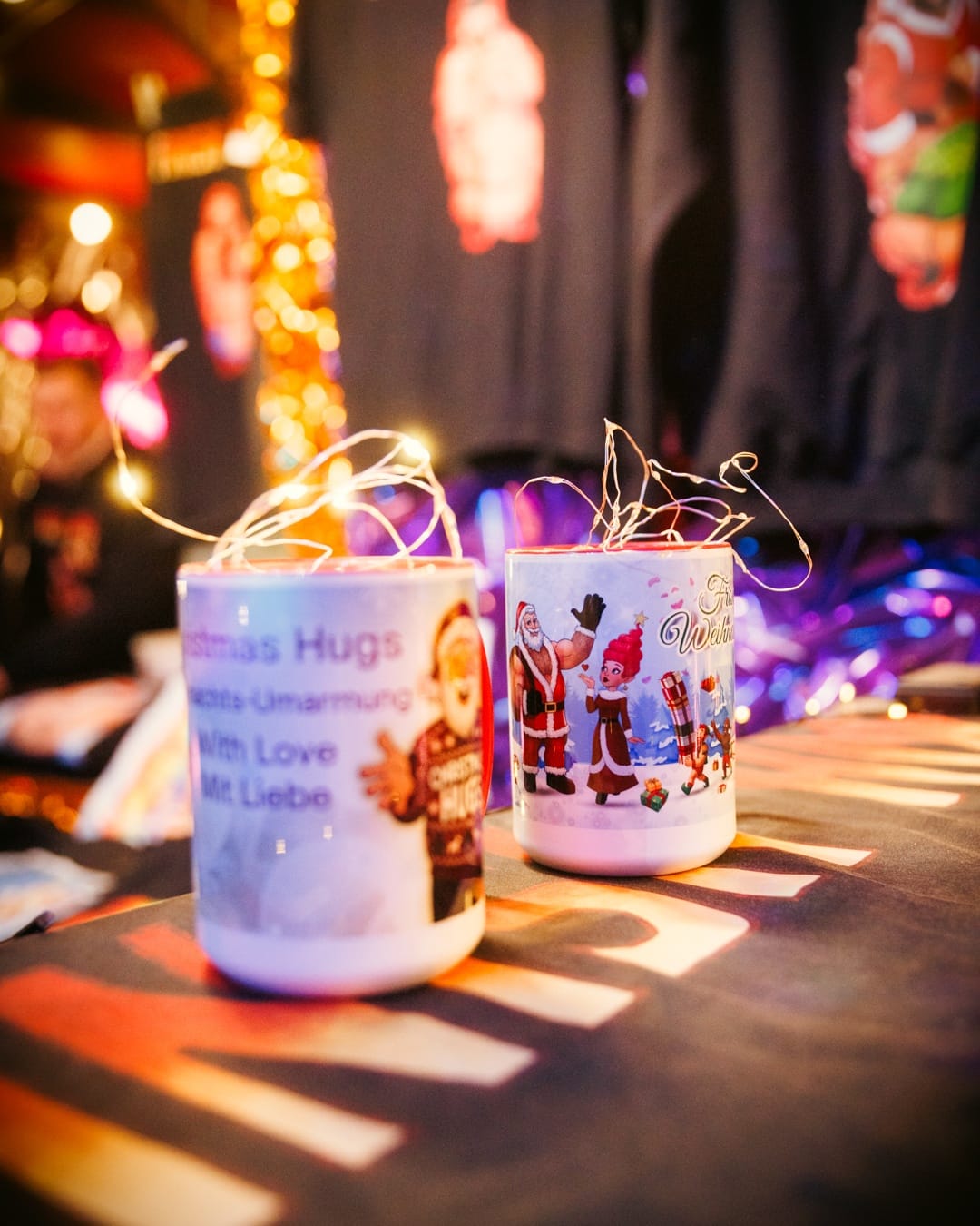 Zwei Tassen mit Weihnachtsmotiven und Lichterkette stehen auf einem Stand mit weihnachtlicher Dekoration im Hintergrund.