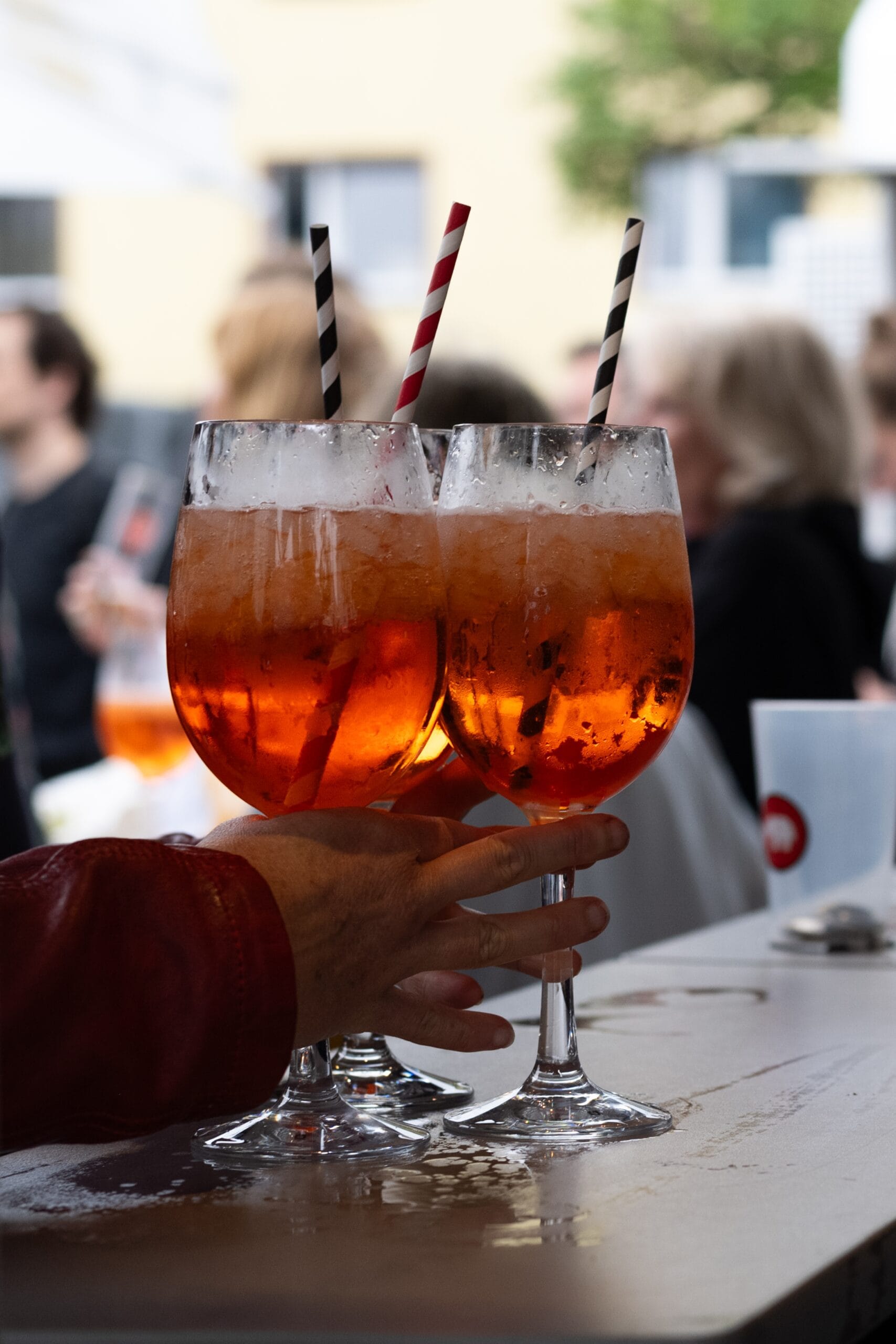 Eine Person hält drei gefüllte Aperol-Gläser mit Eis und Strohhalmen auf einem Tisch im Freien.