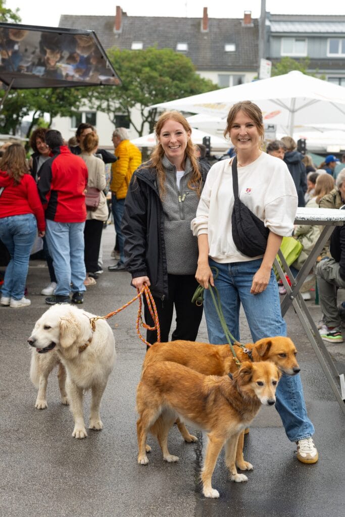 Zwei lächelnde Frauen stehen mit drei Hunden auf einem belebten Platz im Freien, möglicherweise ein Markt.