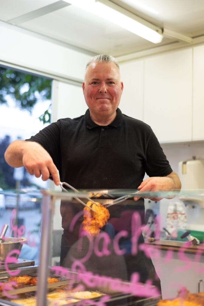 Ein lächelnder Verkäufer in einem Imbisswagen serviert frittierte Speisen mit einer Zange hinter einer gläsernen Theke.