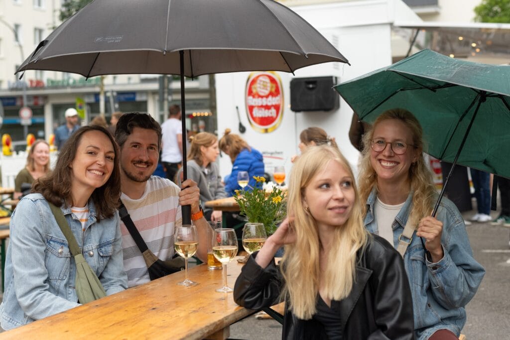Auf einem Feierabendmarkt sitzen Gäste unter Regenschirmen an Biertischen und genießen Wein und die Gesellschaft.