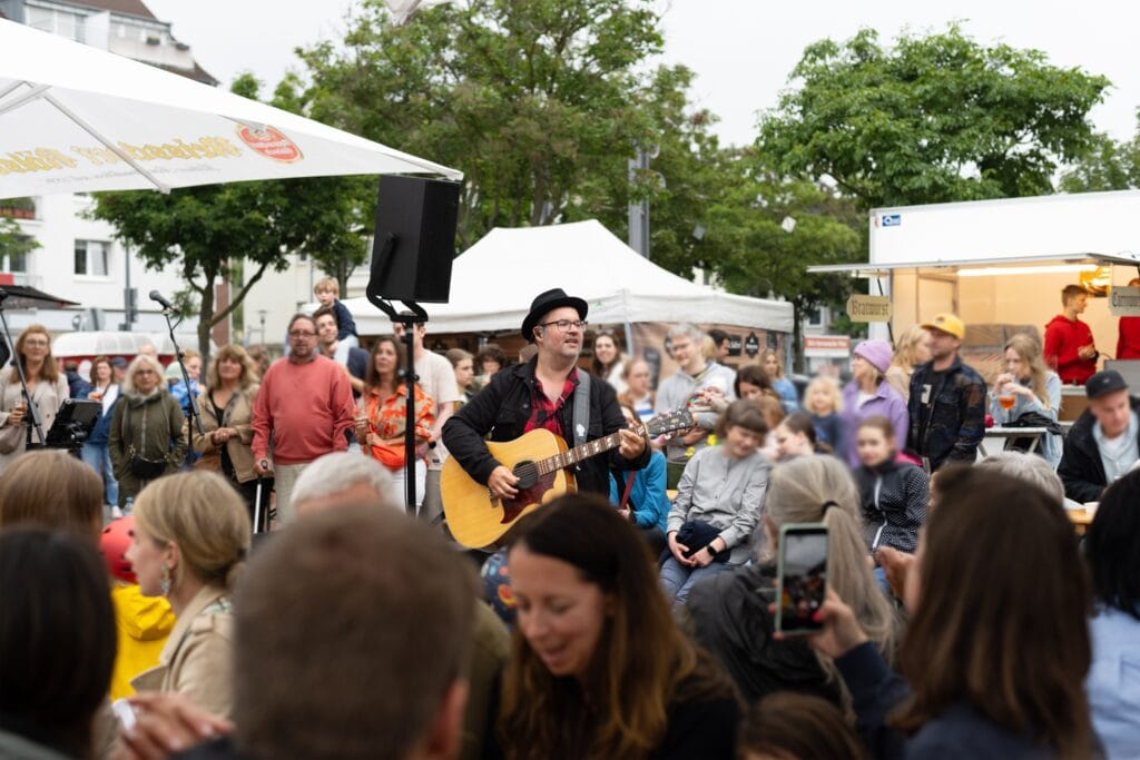 Ein Gitarrenspieler tritt vor einem aufmerksamen Publikum auf, das auf einem Platz mit Imbissständen sitzt.