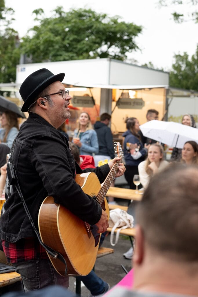 Ein Gitarrist mit Hut singt vor einem Publikum, das an Holztischen sitzt und zuhört.
