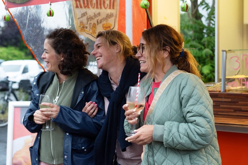 Drei gut gelaunte Frauen stehen zusammen, halten Gläser mit Getränken und lachen herzlich miteinander vor einem Imbissstand.