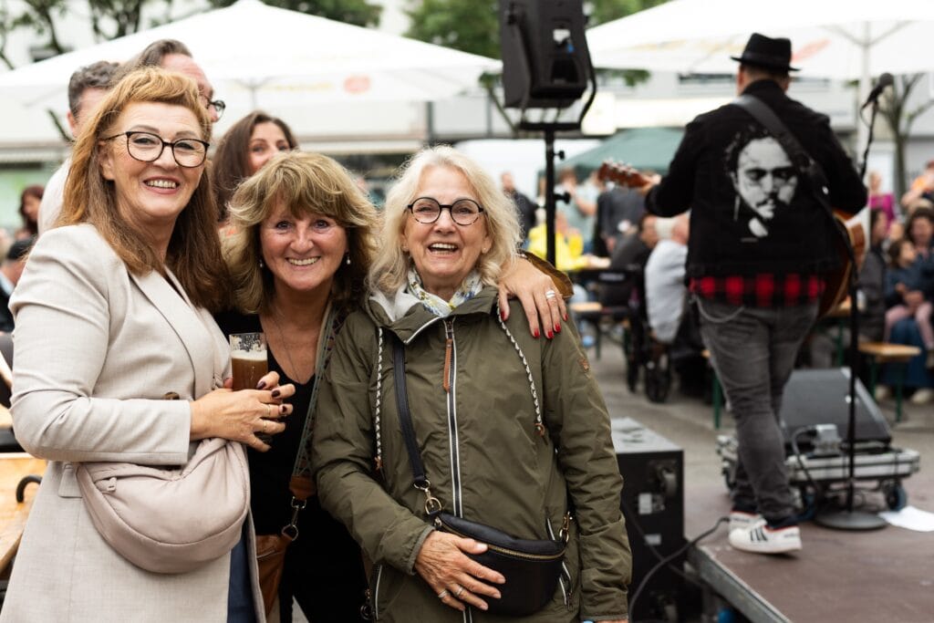 Drei lachende Frauen posieren für ein Foto, während ein Musiker auf der Bühne im Hintergrund spielt.