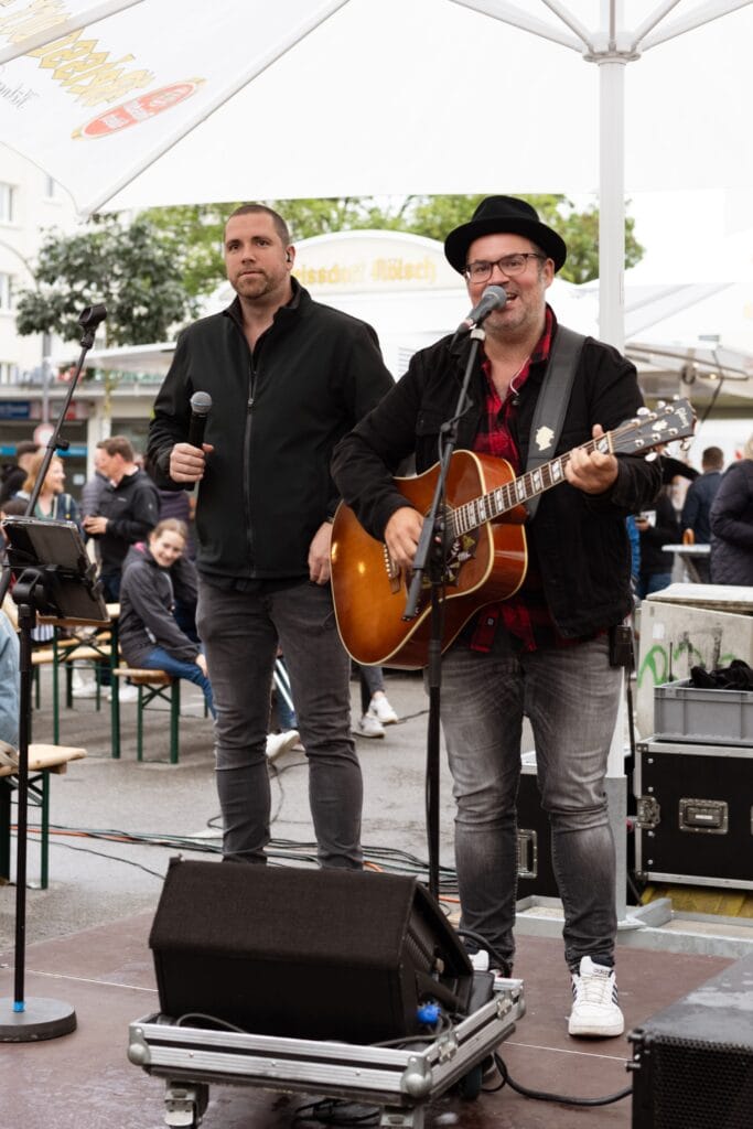 Ein Musikduo spielt live auf einer Bühne: Ein Sänger mit Gitarre und ein Sänger mit Mikrofon.