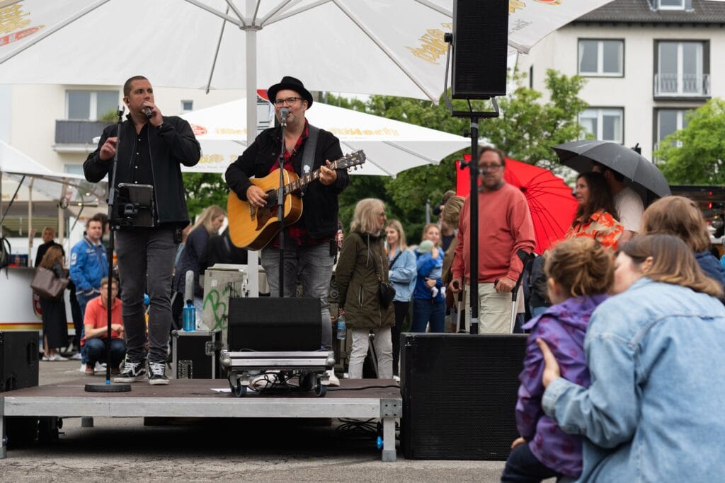 Zwei Musiker performen live auf einer kleinen Bühne vor Publikum, einige Besucher hören mit Regenschirmen zu.