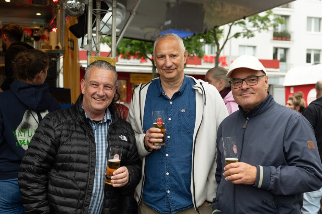 Drei gut gelaunte Männer halten Biergläser in der Hand und lächeln auf einem belebten Platz in die Kamera.