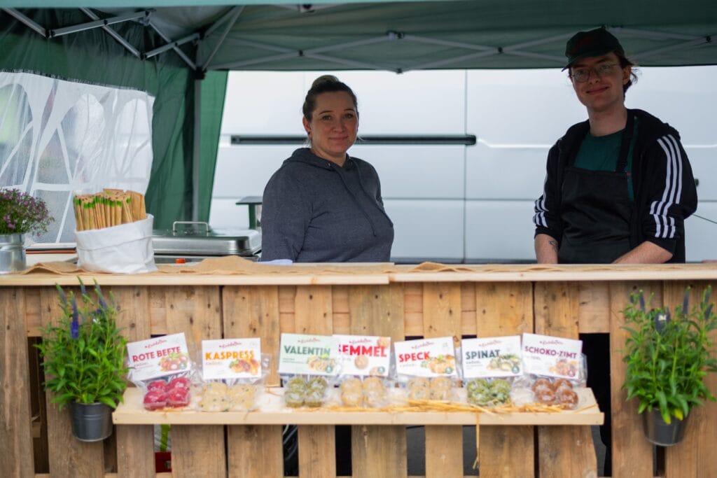 Zwei freundliche Verkäufer stehen an einem Marktstand aus Holzpaletten und präsentieren verschiedene Sorten von Lebensmitteln in Verpackungen.