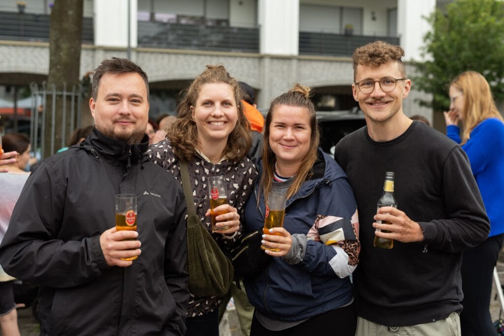 Vier gut gelaunte Personen, zwei Frauen und zwei Männer, halten Biergläser und eine Flasche in den Händen und lachen.
