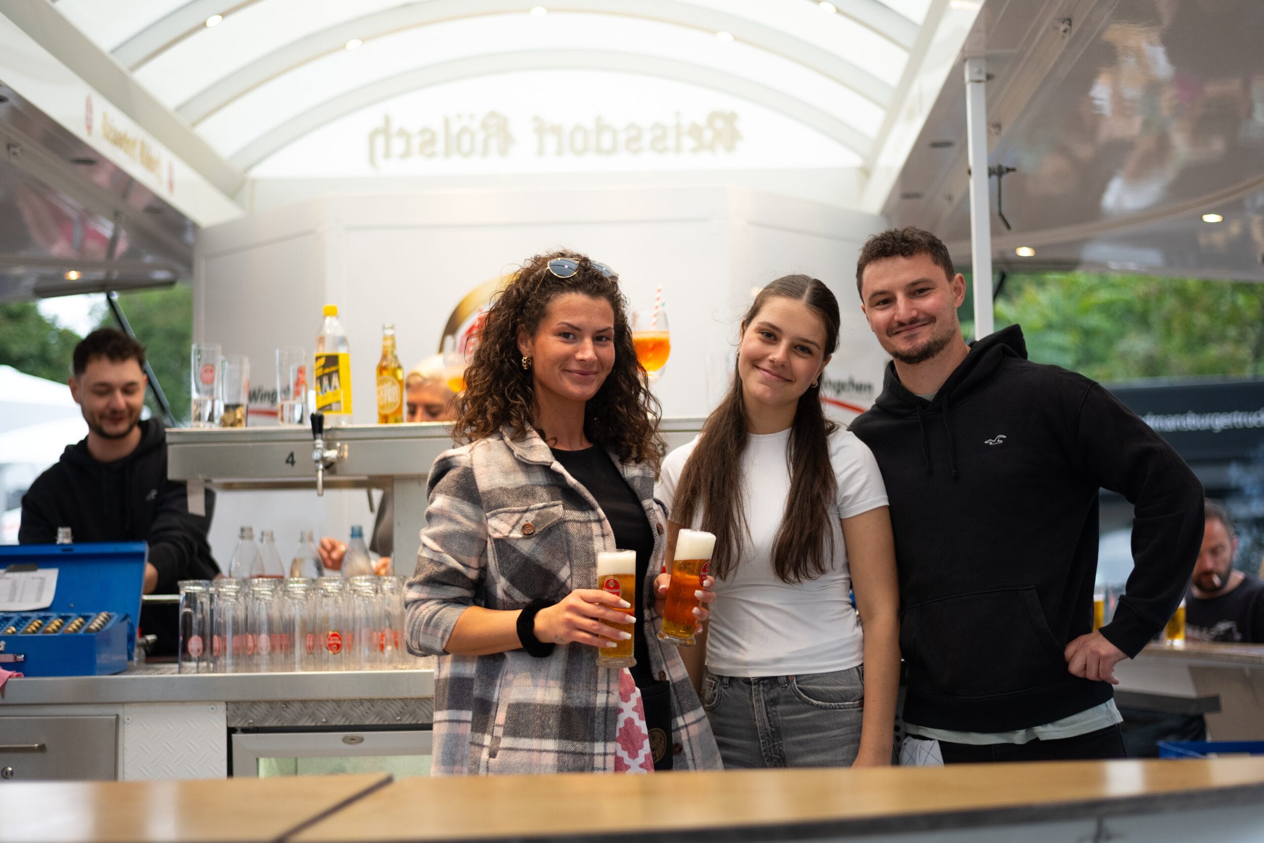 Zwei junge Frauen und ein Mann halten Biergläser in der Hand und lächeln freundlich in die Kamera.