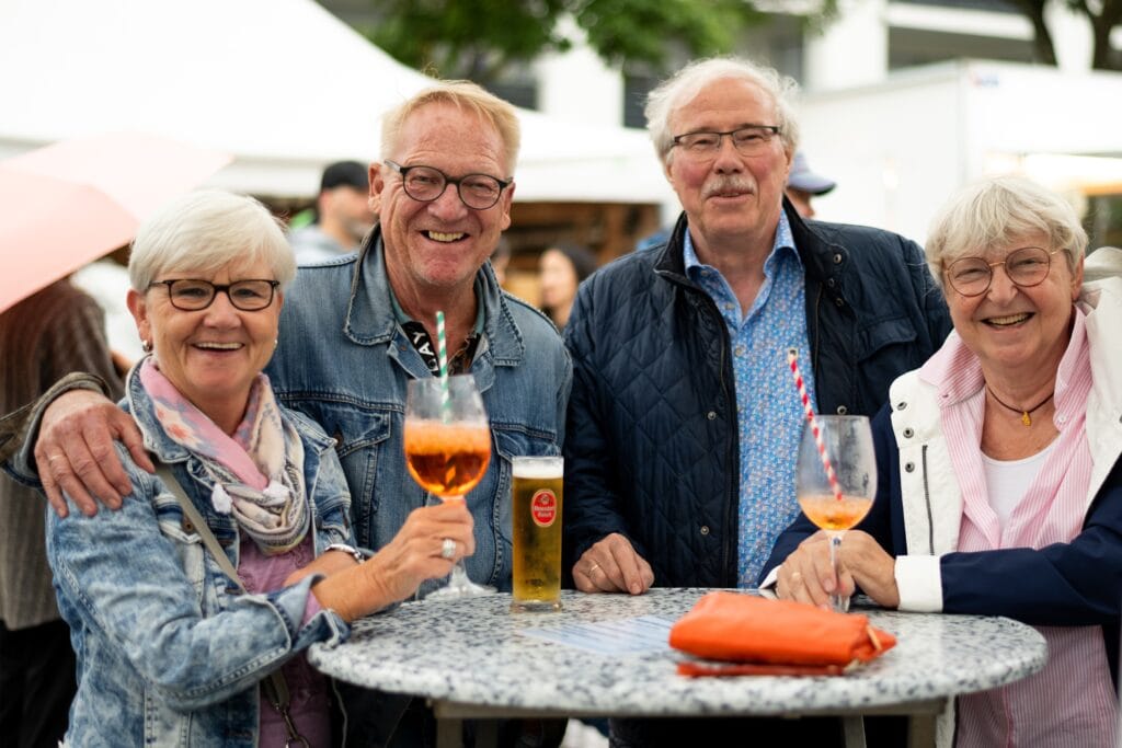 Zwei Paare mittleren Alters stehen an einem Tisch und genießen ihre Getränke im Freien, lachend und gut gelaunt.