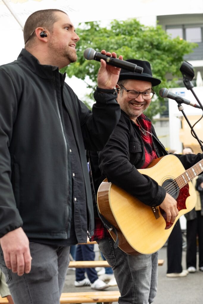 Zwei Musiker stehen auf einer Bühne und singen in Mikrofone, einer spielt Gitarre, beide tragen lässige Kleidung.