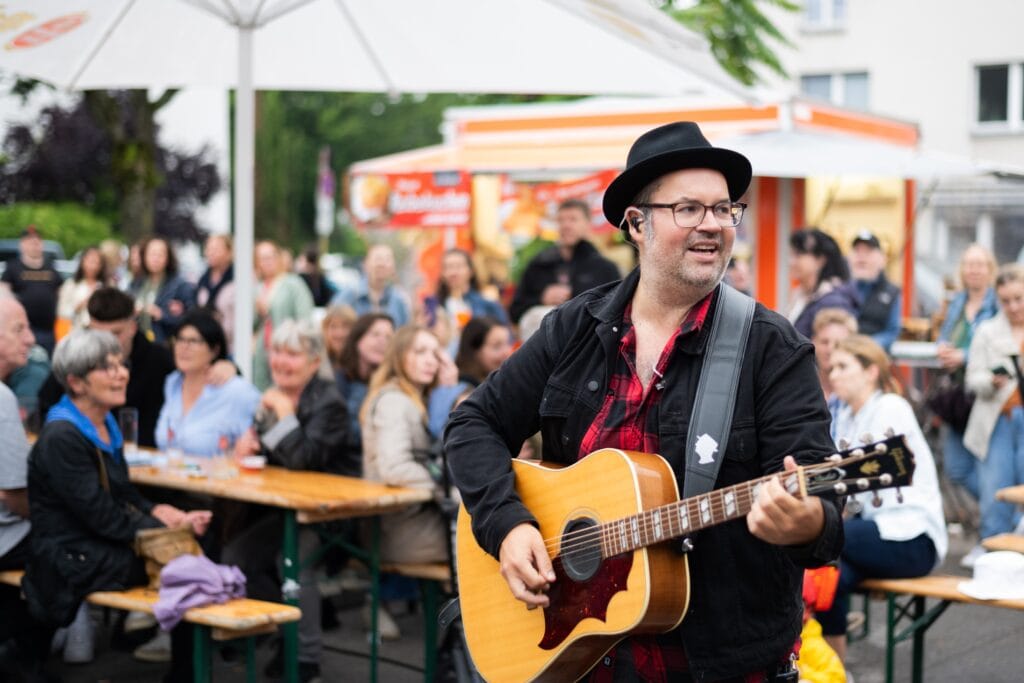 Ein Musiker mit Hut spielt Gitarre vor einem aufmerksam lauschenden Publikum im Freien.