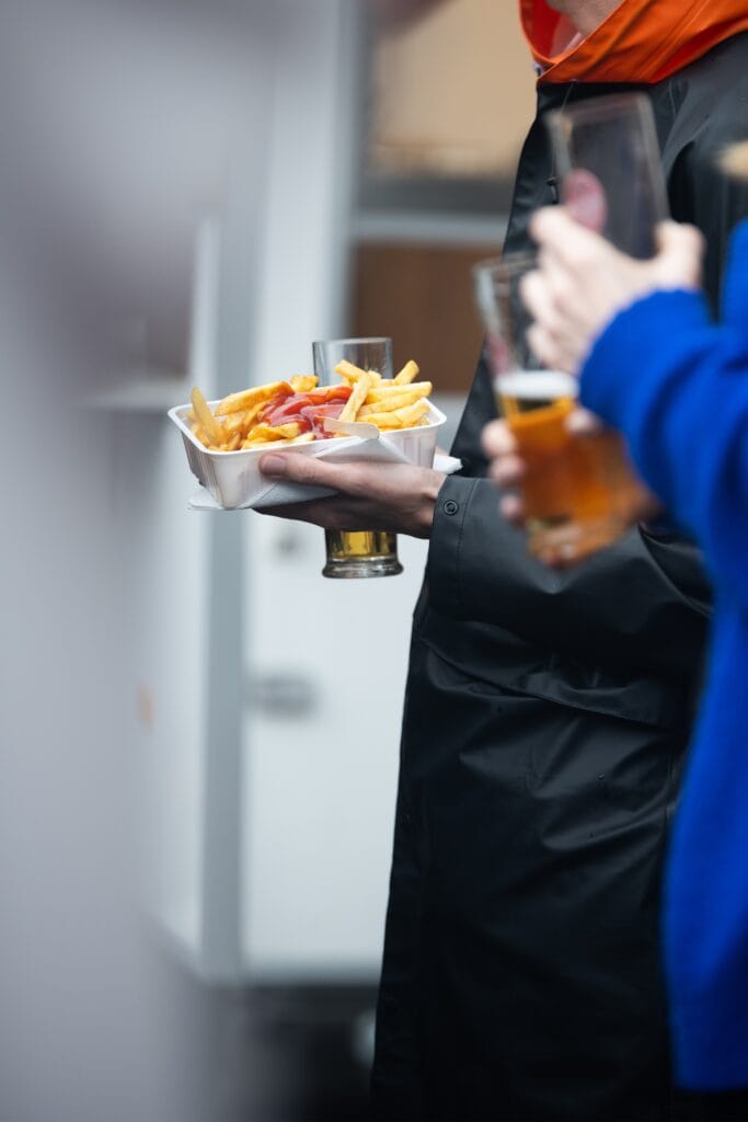 Eine Person hält Pommes mit Ketchup und ein Bier in der Hand auf einem Event.