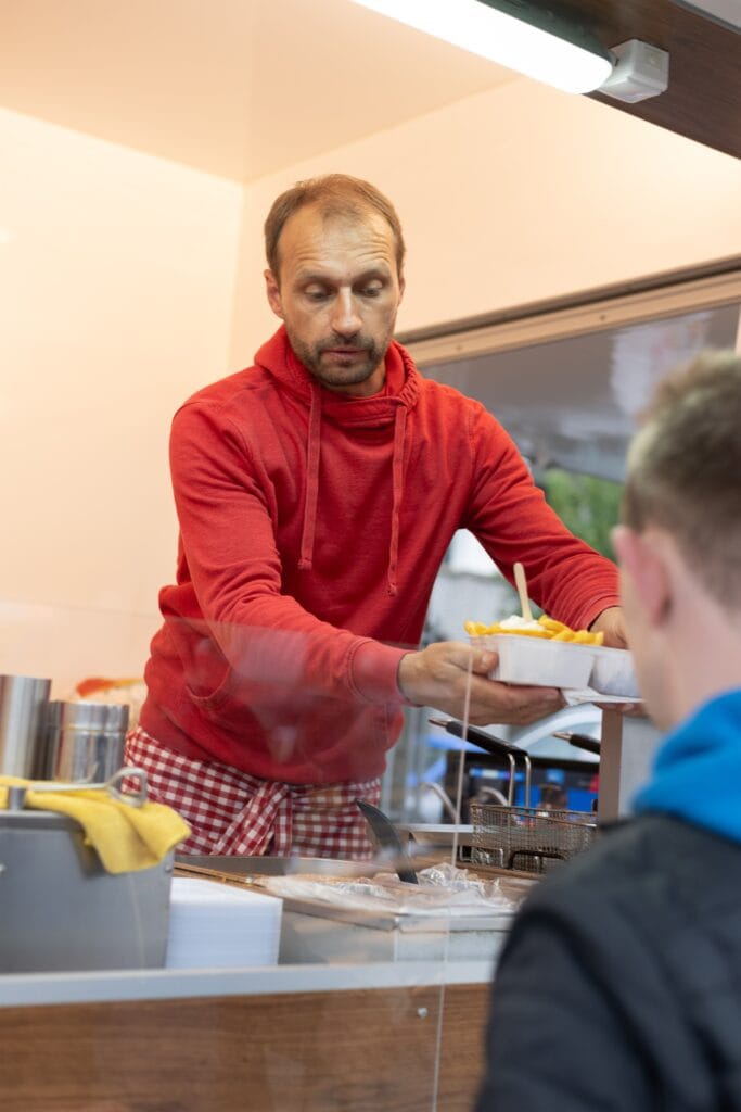 Ein Verkäufer in rotem Hoodie reicht einem Kunden eine Portion Pommes Frites mit Mayonnaise in einer weißen Schale.
