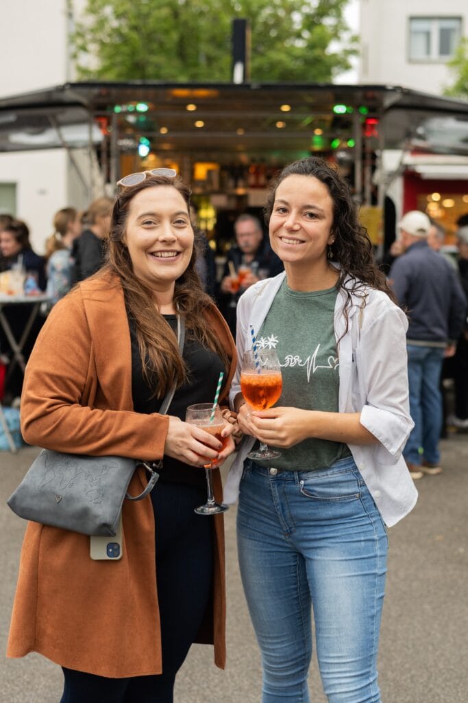 Zwei lächelnde Frauen mit Getränken stehen vor einem Imbisswagen auf einem Event und genießen den Abend.