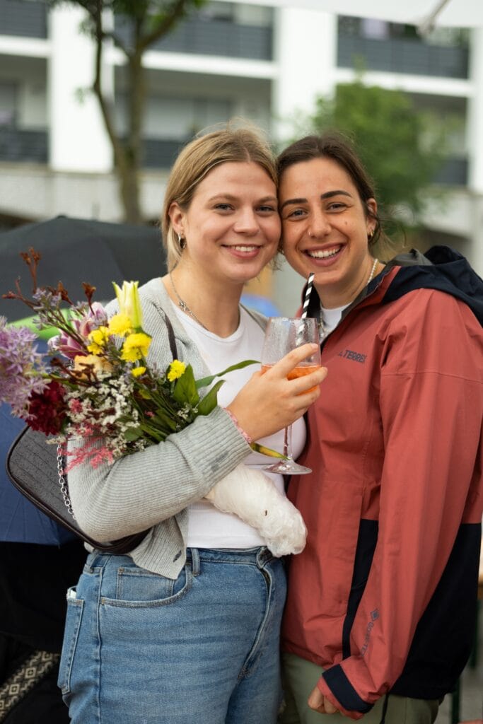 Zwei junge, lächelnde Frauen posieren mit Blumenstrauß und Getränk auf einer Veranstaltung im Freien für ein Foto.