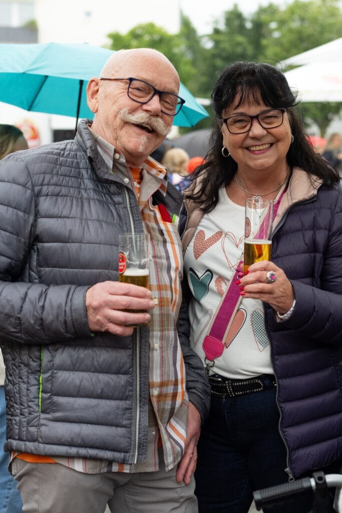 Ein freundliches Paar mit Brillen und Jacken prostet mit Biergläsern auf einem Event im Freien zu.
