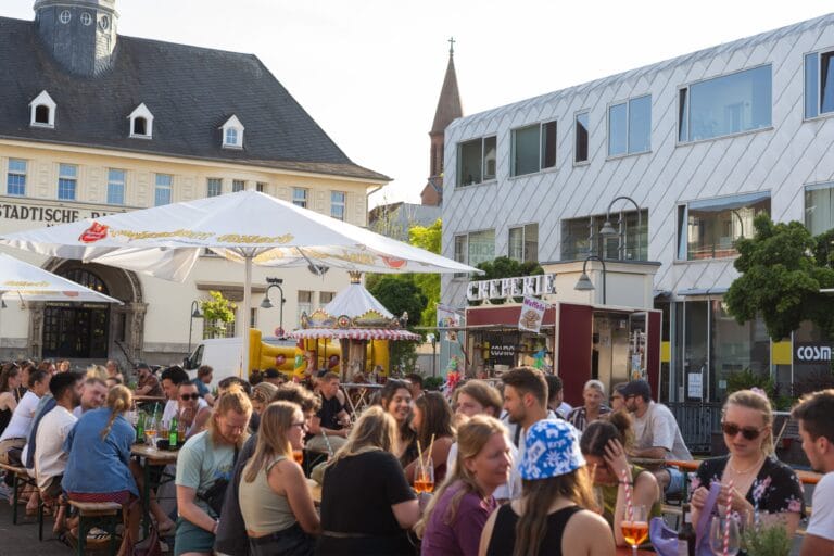 Auf dem Feierabendmarkt in Köln sitzen und stehen viele junge Menschen an Biertischen und genießen Getränke bei Sonnenschein.