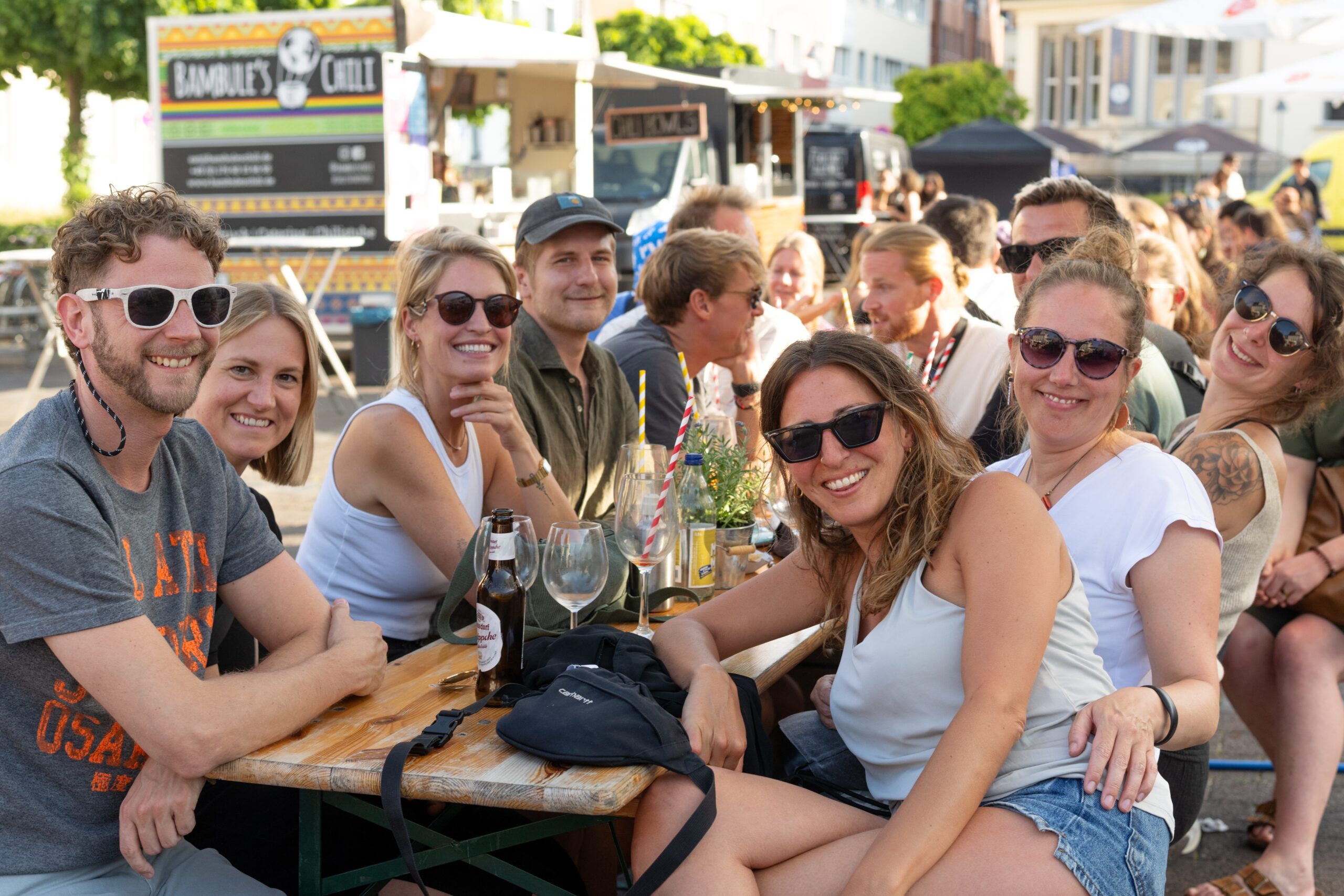 Eine Gruppe gut gelaunter junger Menschen sitzt an einem Tisch auf einem Event im Freien und lächelt in die Kamera.