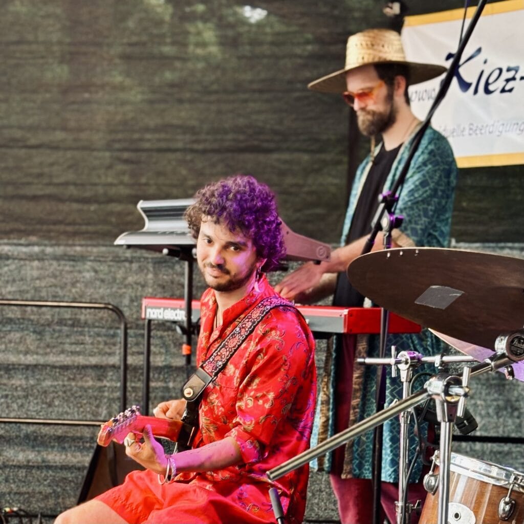 Zwei Musiker spielen auf einer Bühne, ein Gitarrist in rotem Hemd schaut in die Kamera, Keyboarder mit Hut im Hintergrund.