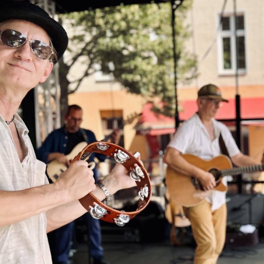 Eine Band spielt live auf einer Bühne, ein Mann mit Hut und Tamburin im Vordergrund, Gitarrist und Bassist dahinter.