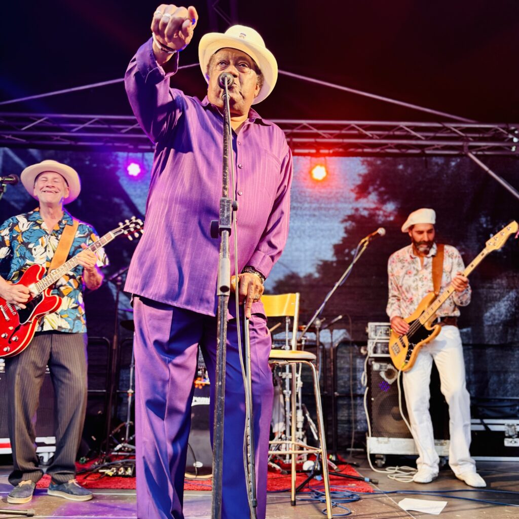 Ein Sänger in lila Anzug steht mit zwei Gitarristen auf der Bühne und performt.