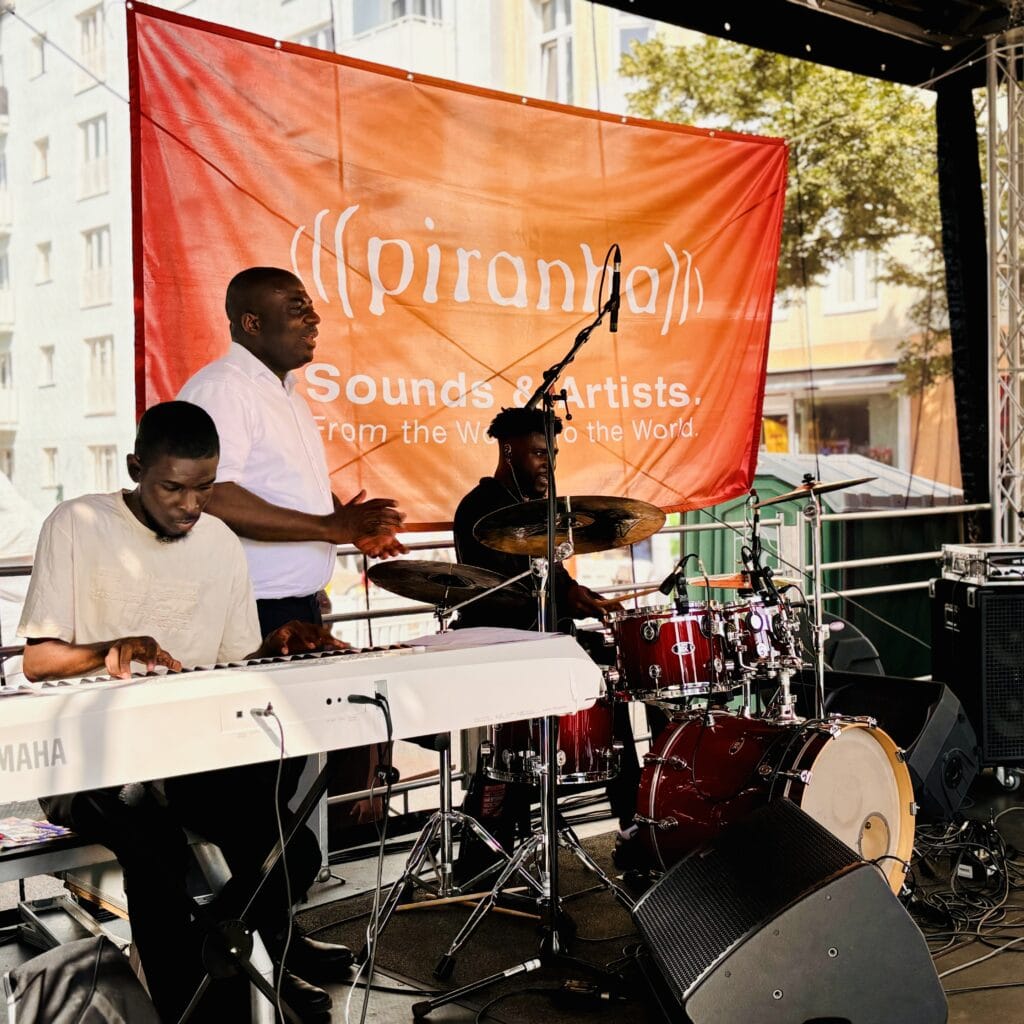 Drei Musiker spielen auf einer Bühne vor einem großen, orangefarbenen Banner mit dem Schriftzug "piranha Sounds & Artists."