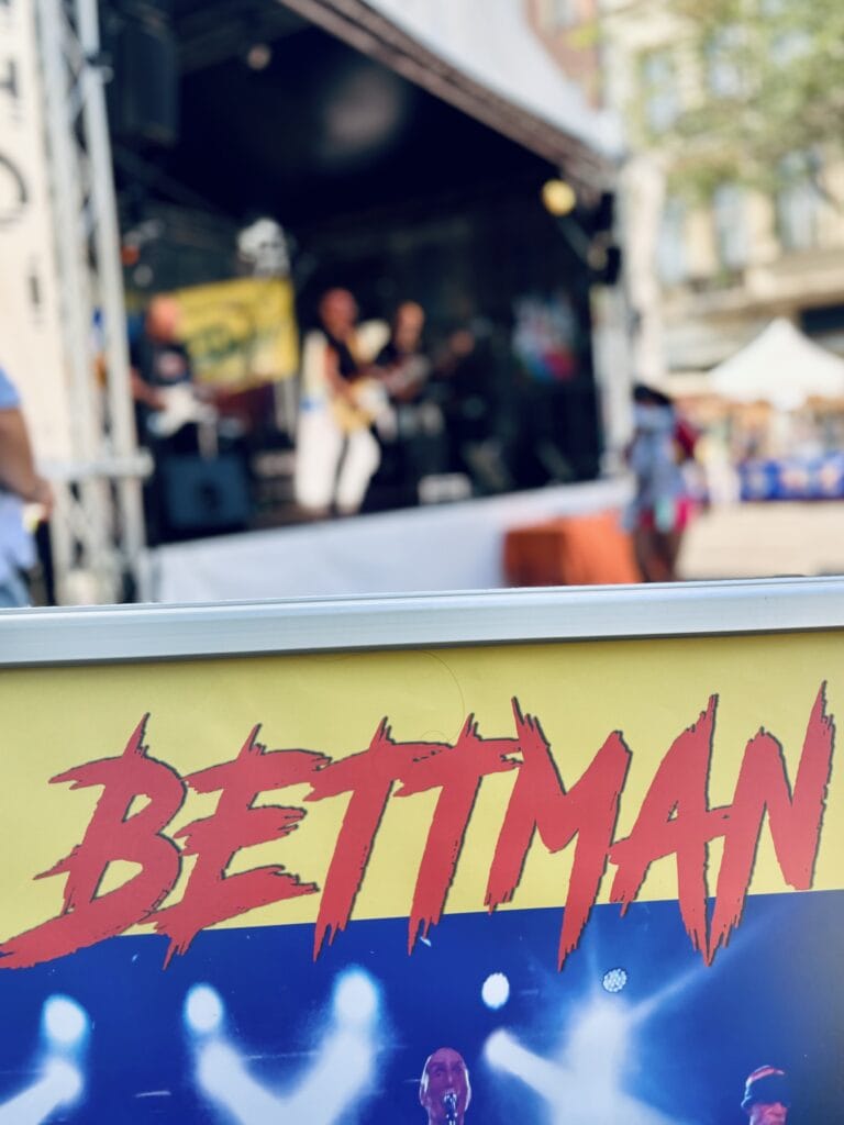 Vor einer Bühne mit Musikern steht ein Poster mit dem Schriftzug "Bettman" in roter Farbe und gelbem Hintergrund.