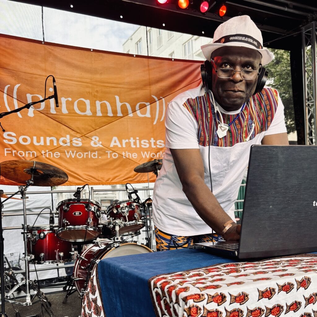 Ein DJ mit Hut und Kopfhörern legt Musik auf, ein Schlagzeug und Banner sind im Hintergrund zu sehen.