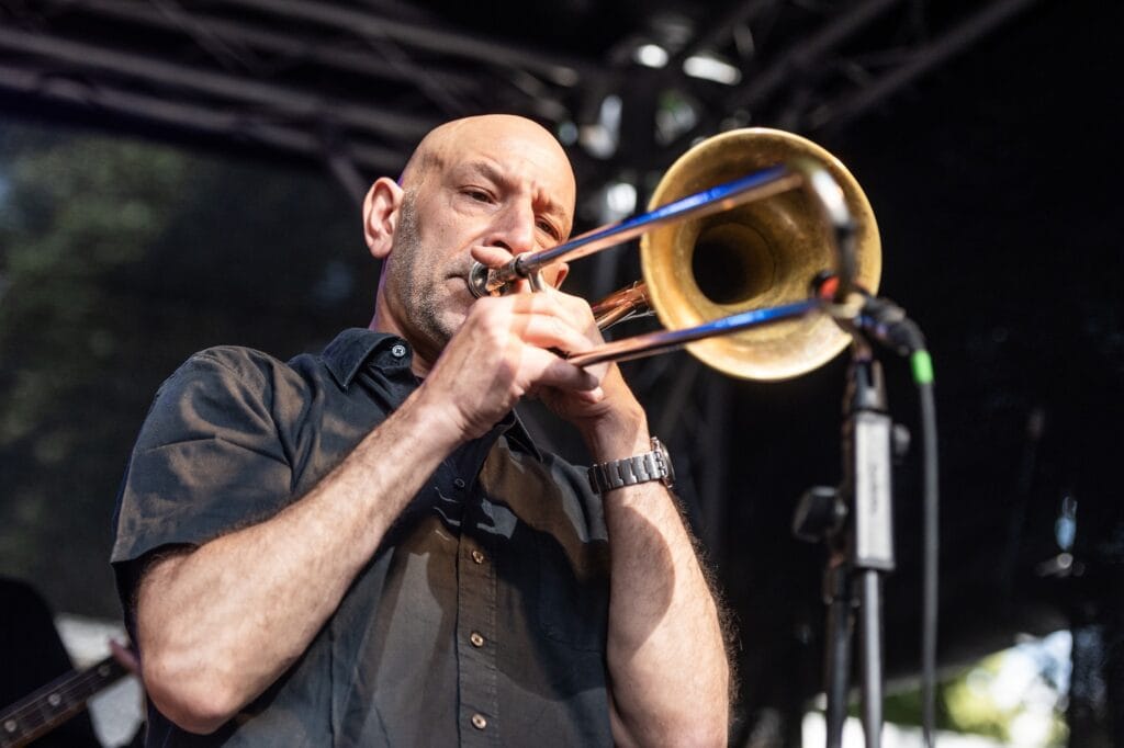 Ein Mann mit Glatze und Bart spielt auf einem Festival in Berlin Kreuzberg leidenschaftlich Posaune, während er in ein Mikrofon singt und dabei ein schwarzes Hemd und eine Armbanduhr trägt.