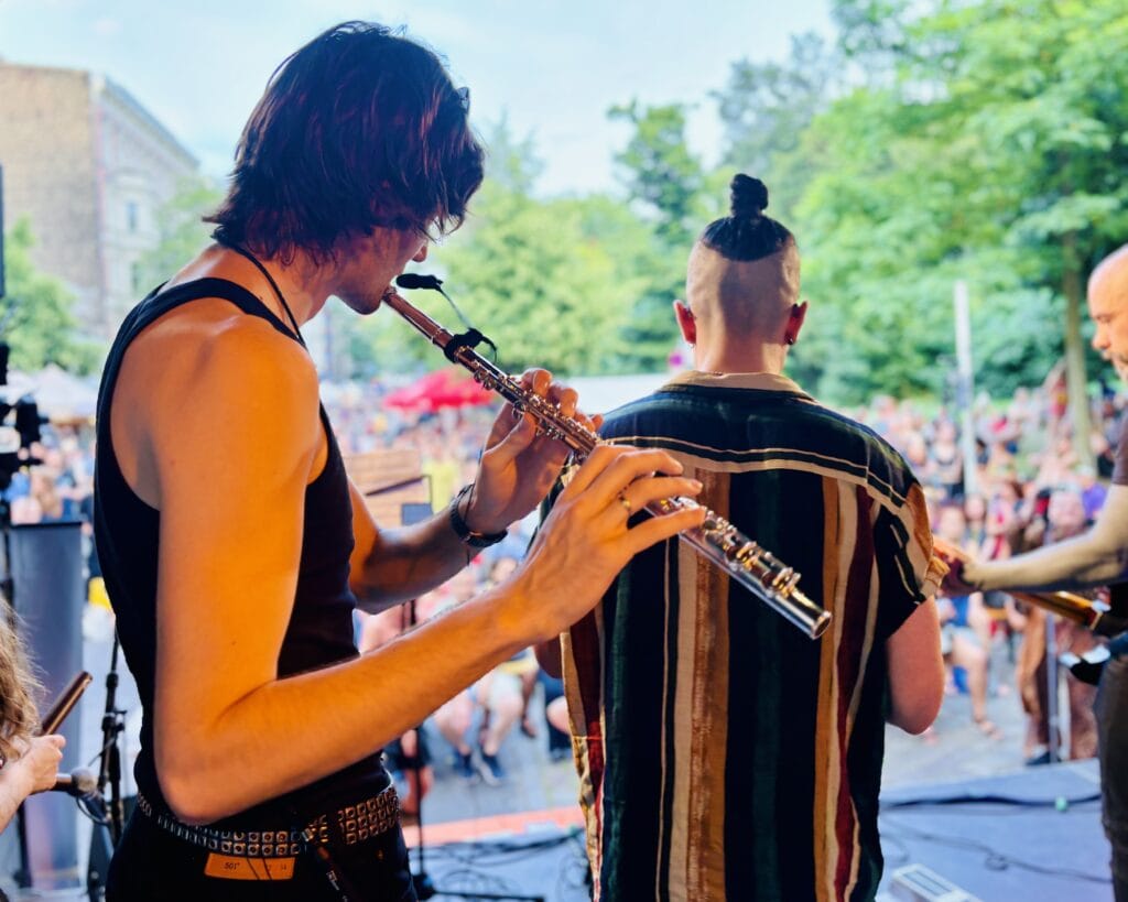 Ein junger Mann mit langen Haaren spielt auf einer Bühne Flöte, im Hintergrund andere Musiker und ein Publikum.