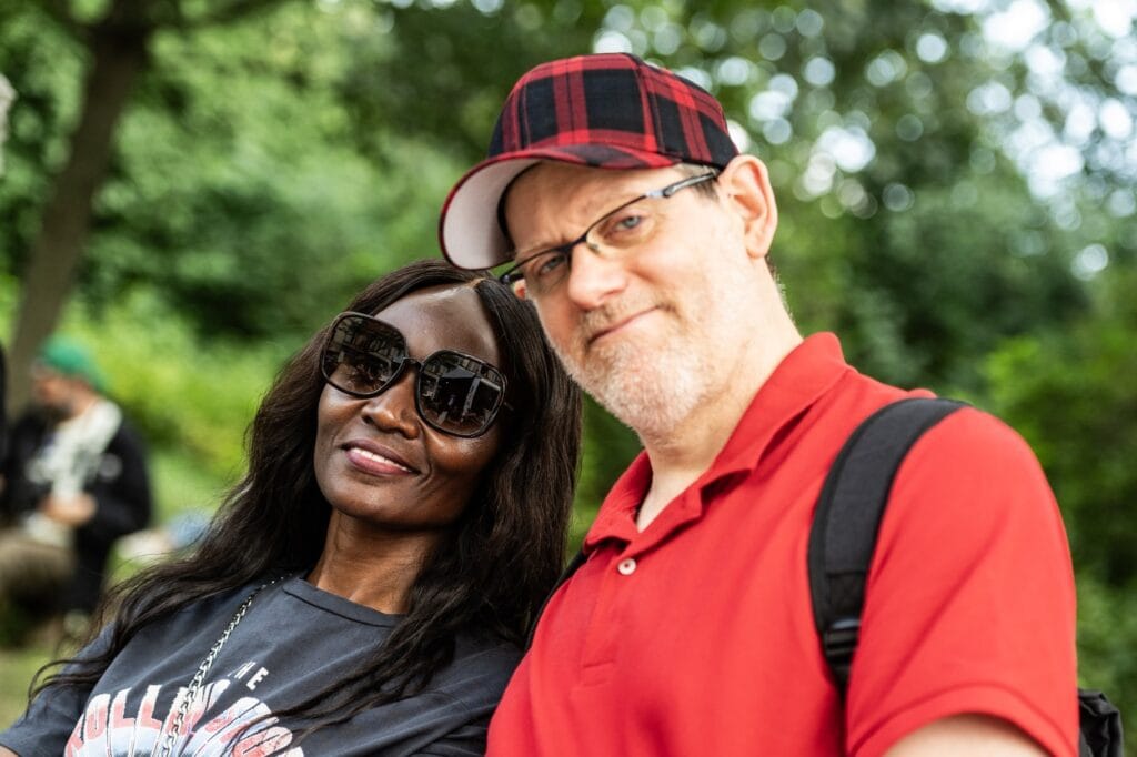 Ein lächelndes Paar, Frau mit Sonnenbrille und Mann mit karierter Kappe, posiert entspannt vor grünem Hintergrund im Freien.
