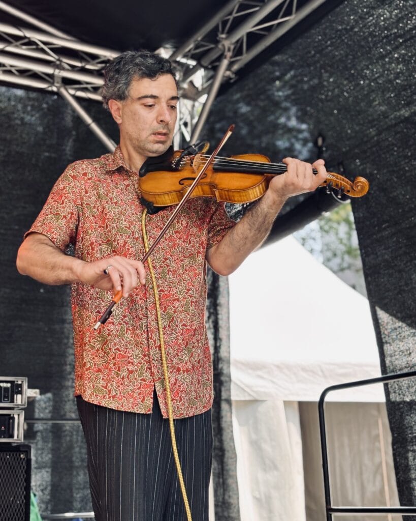 Ein Mann mit gemustertem Hemd spielt auf einer Bühne eine Geige und tritt bei einem Event auf.