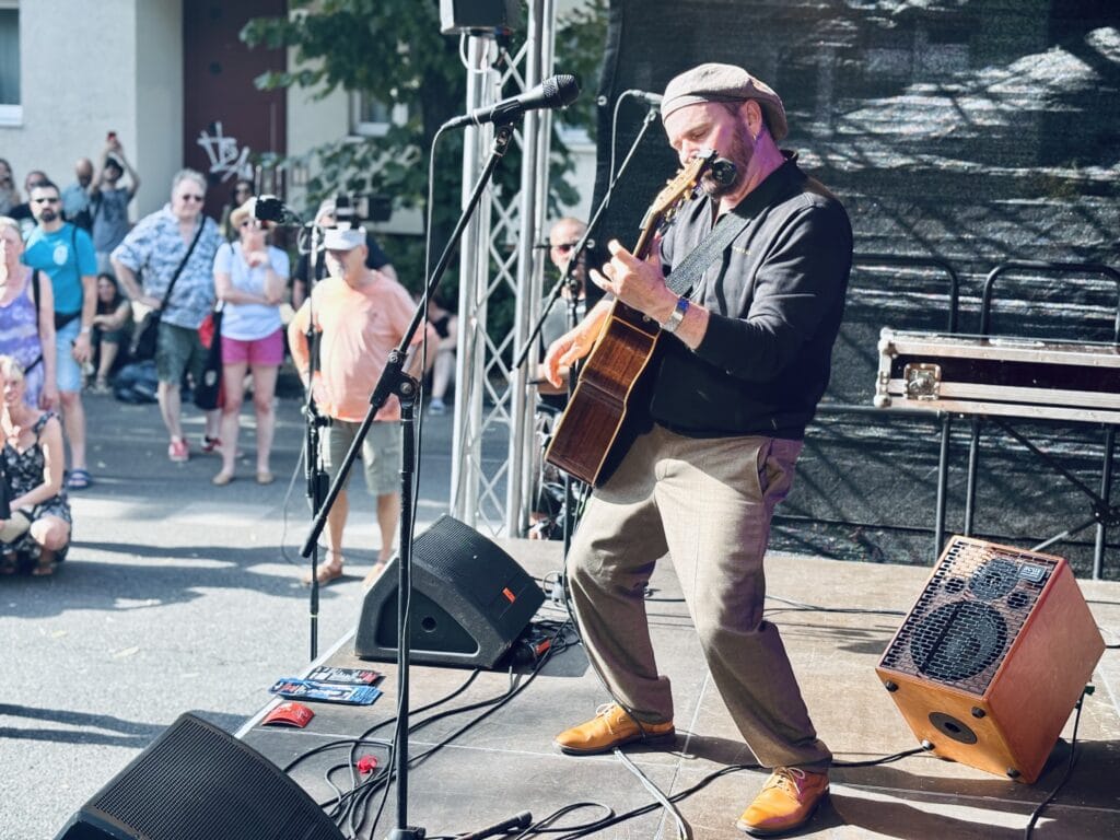 Ein Musiker mit Gitarre und Mundharmonika spielt auf einer Bühne vor Publikum im Freien.