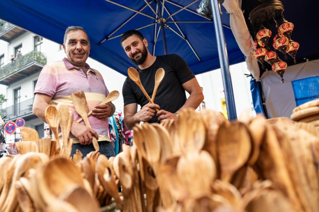 Zwei Männer präsentieren stolz ihre Holzlöffel auf einem Marktstand unter einem blauen Sonnenschirm.