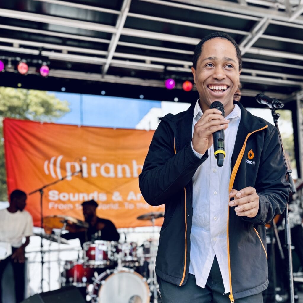 Ein lachender, junger Mann mit Mikrofon steht auf einer Bühne vor einem Banner und einer Band im Hintergrund.