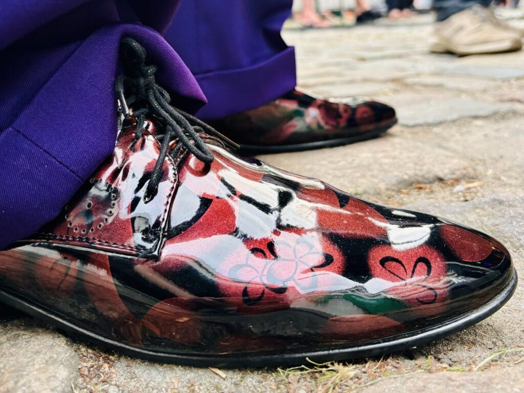 Ein Paar glänzende Lackschuhe mit rotem Blumenmuster stehen vor lila Hosenbeinen auf einem steinigen Untergrund.