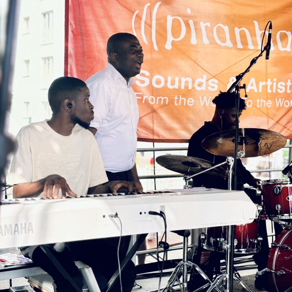 Ein Trio spielt live Musik auf einer Bühne, mit Keyboard, Schlagzeug und einem Sänger vor einem Banner.