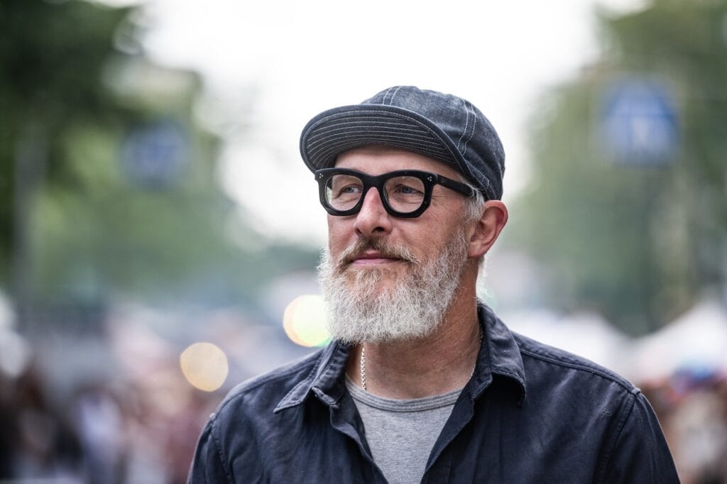 Ein Mann mit grauem Bart, Brille und Kappe schaut aufmerksam auf dem belebten Straßenfest.